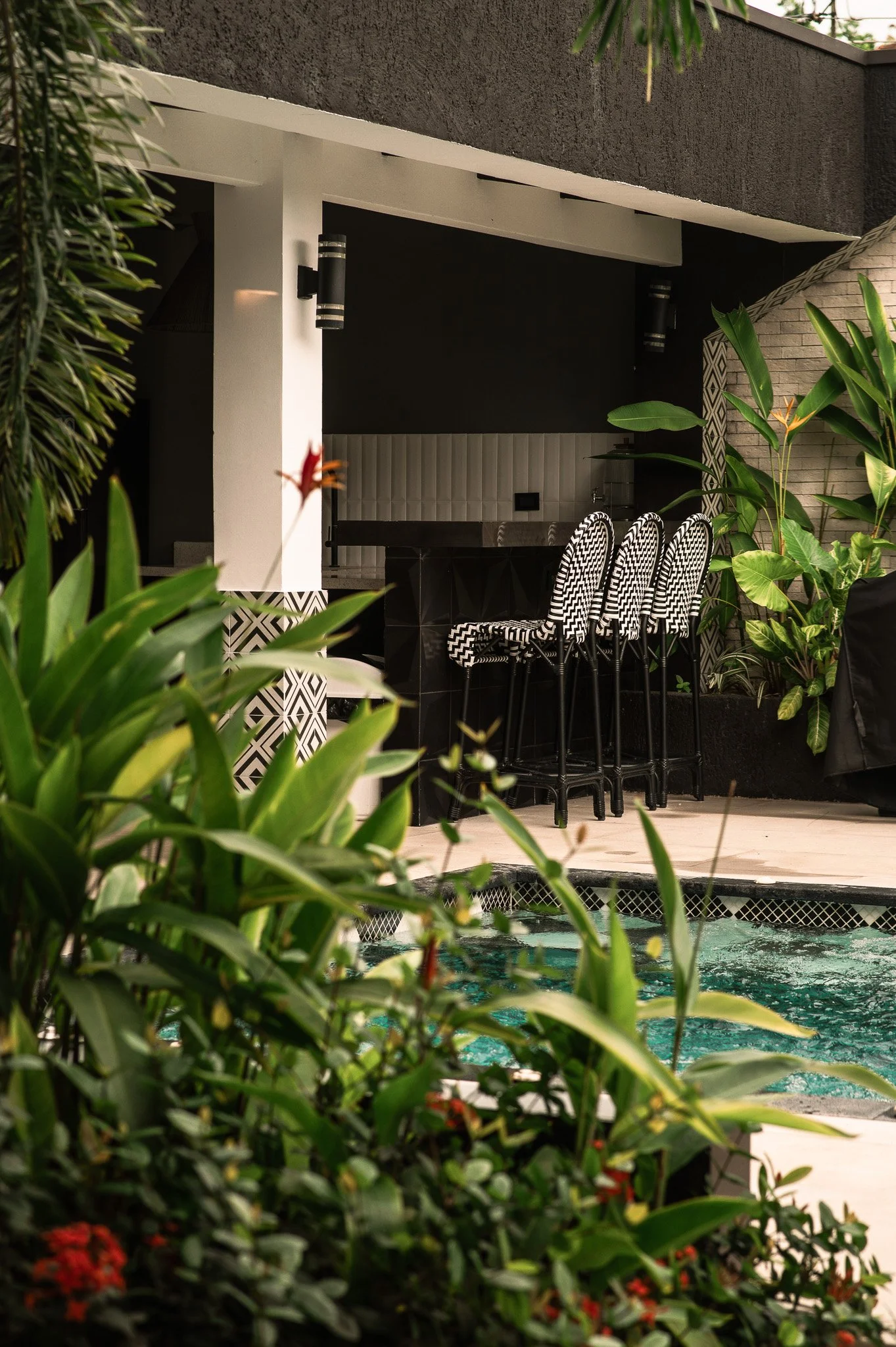 View of an outdoor patio area with a pool, surrounded by lush green tropical plants and outdoor furniture, with a black and white patterned wall and lighting fixtures in the background.
