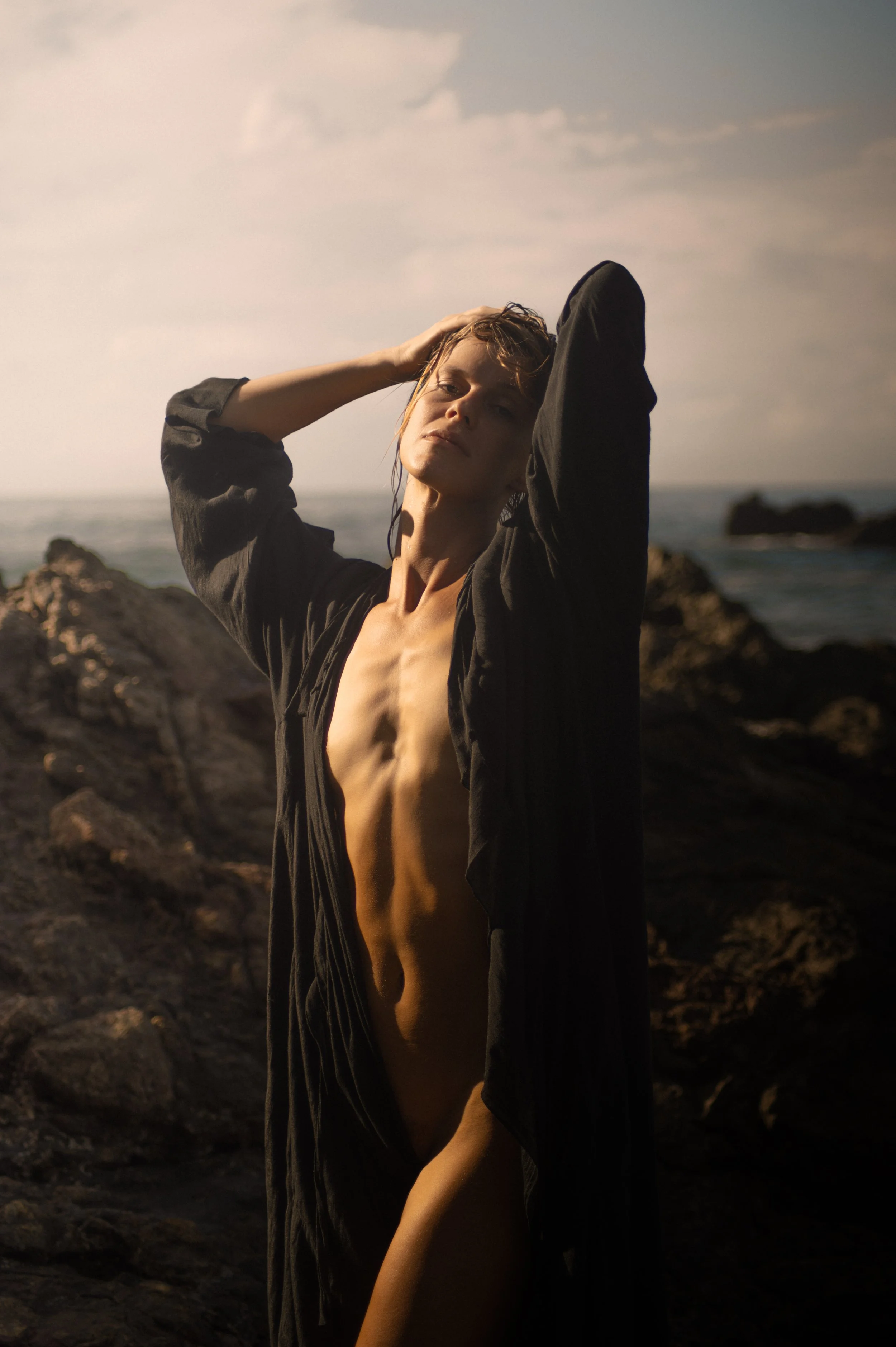 A person with wet hair standing on a rocky shoreline, wearing an open black shirt with no shirt underneath, looking at the camera with one hand in their hair and the other arm across their side, during sunset.
