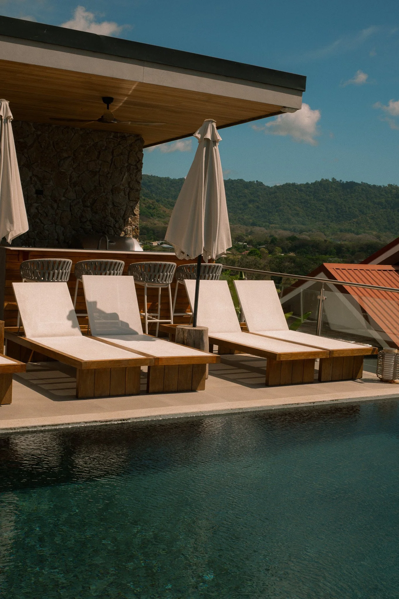 A luxurious outdoor pool area with wooden lounge chairs and white umbrellas, overlooking green hills and a partly cloudy sky.