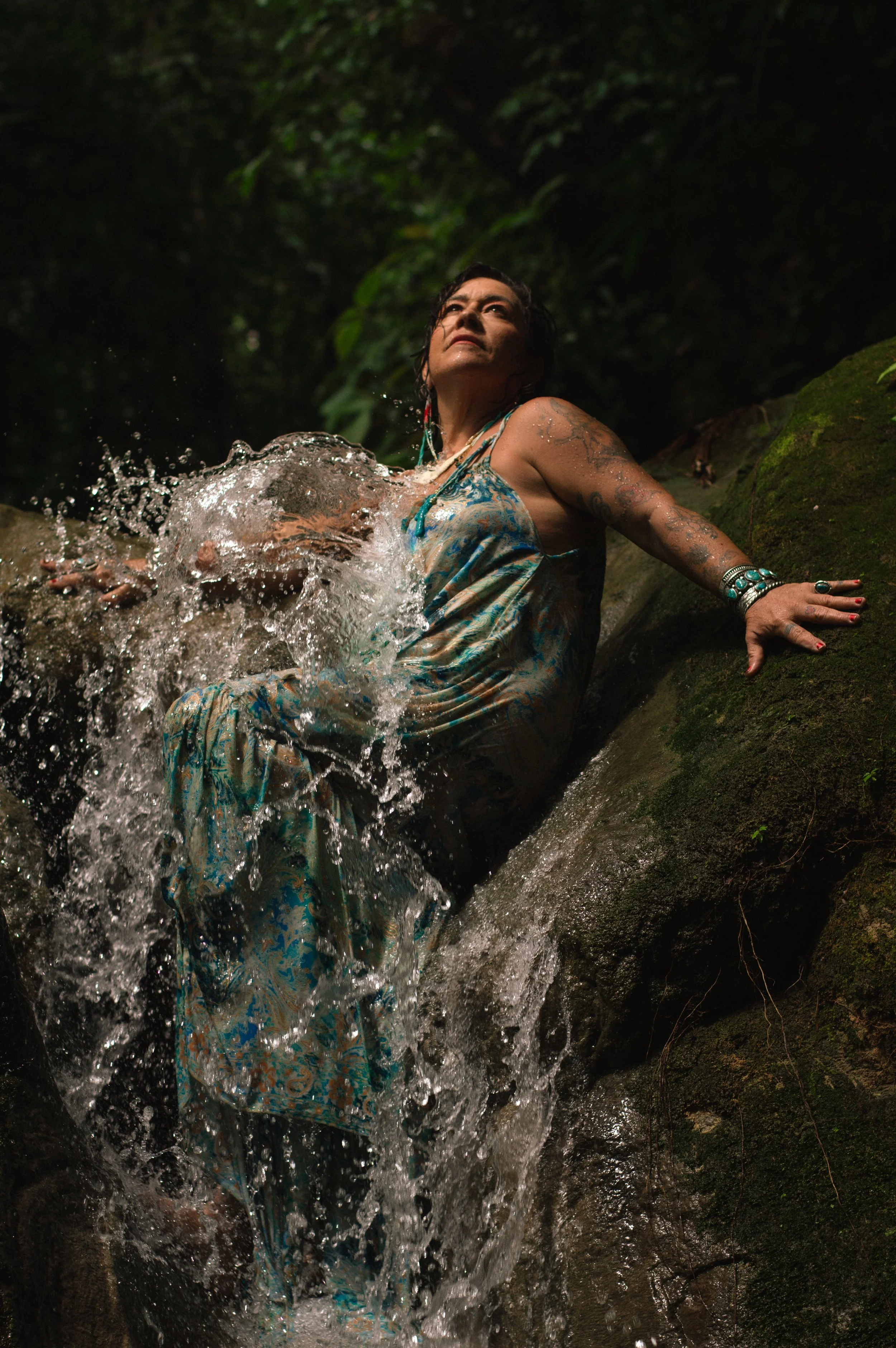 Woman with tattoos and jewelry sitting in a natural water stream surrounded by green foliage at night.