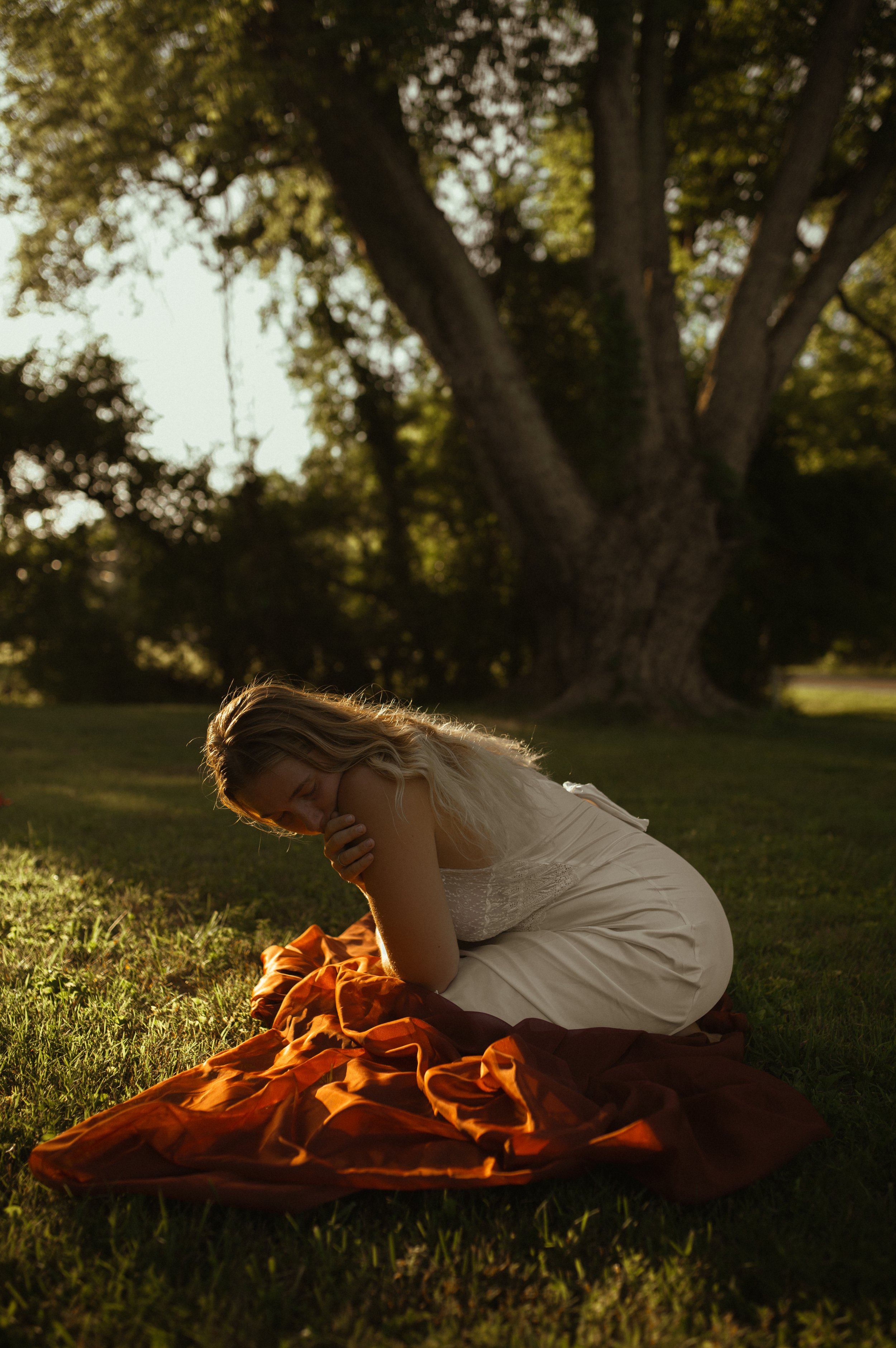 A woman in a white dress sits on the grass, covered by an orange blanket, with her head bowed and hands clasped near her face, in a park during late afternoon or early evening with large trees and sunlight in the background.