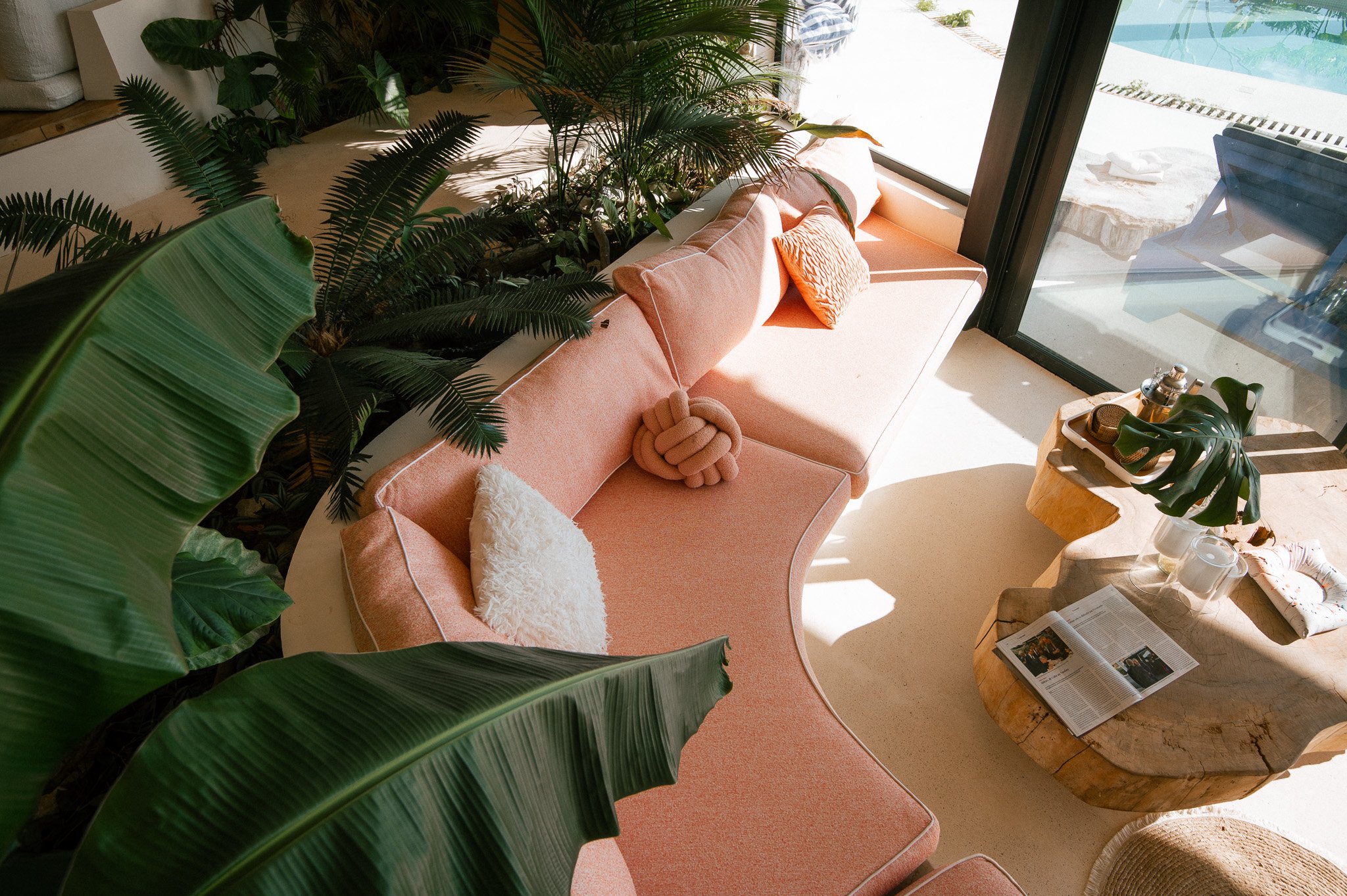 Sunlit pink sofa with a textured orange pillow and a white fuzzy pillow beside a wooden coffee table with a plant, newspaper, and candles, next to a large glass door with a swimming pool outside.