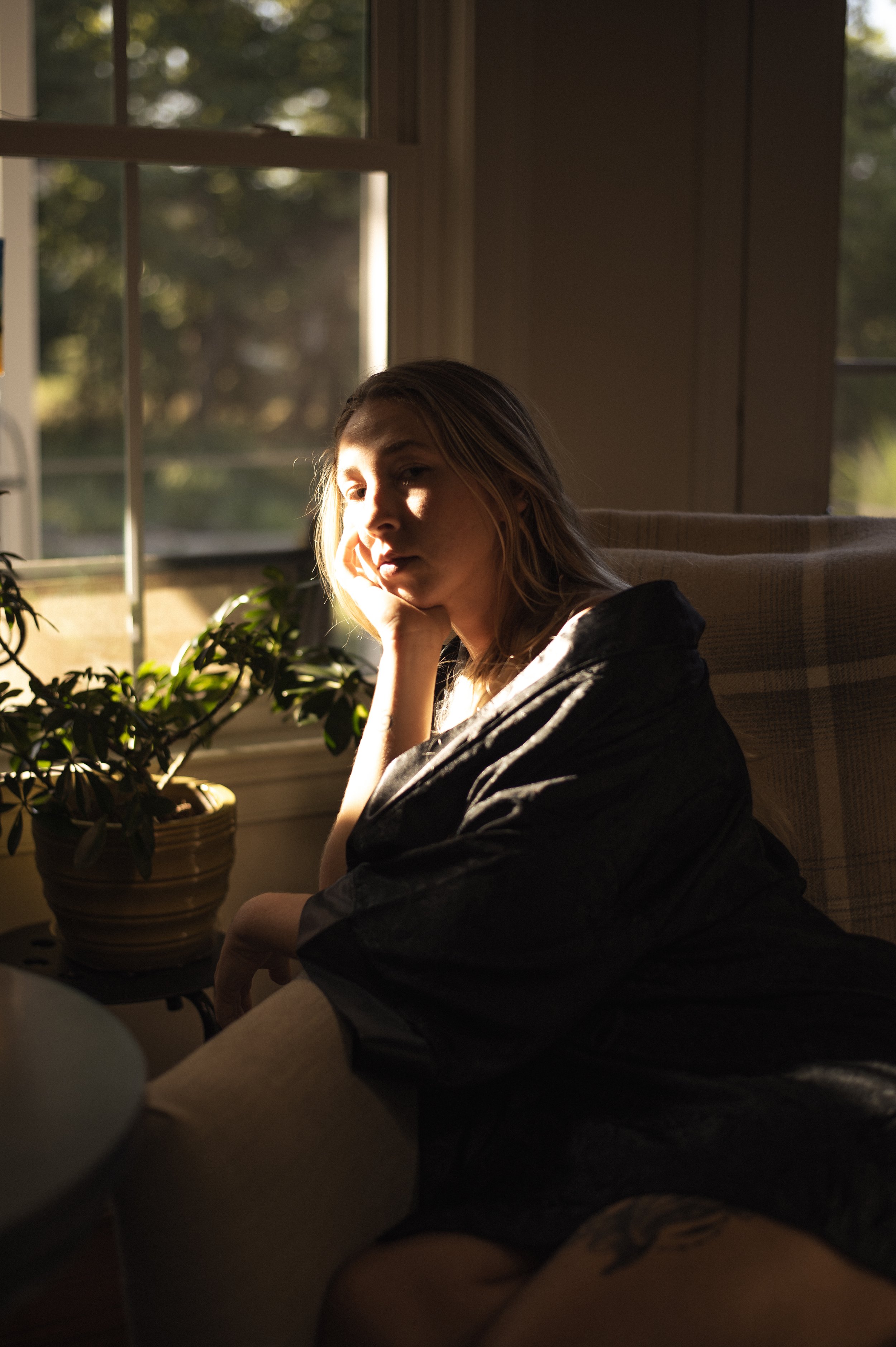 A woman sitting in a sunlit room, resting her chin on her hand, with a potted plant and a window in the background.