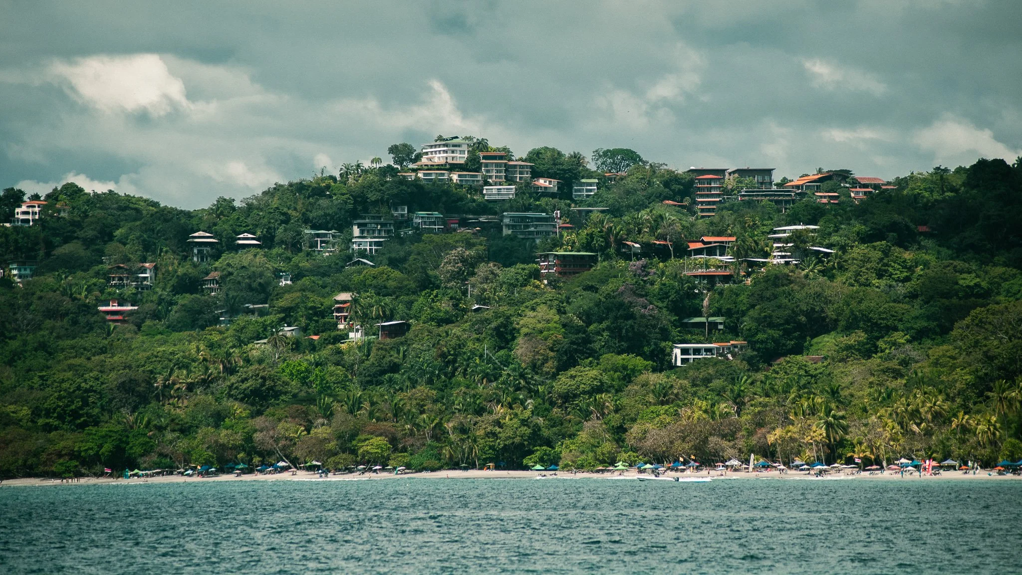 Hillside overlooking a beach with umbrellas and boats, with modern houses and greenery on the hilltop and stormy clouds overhead.