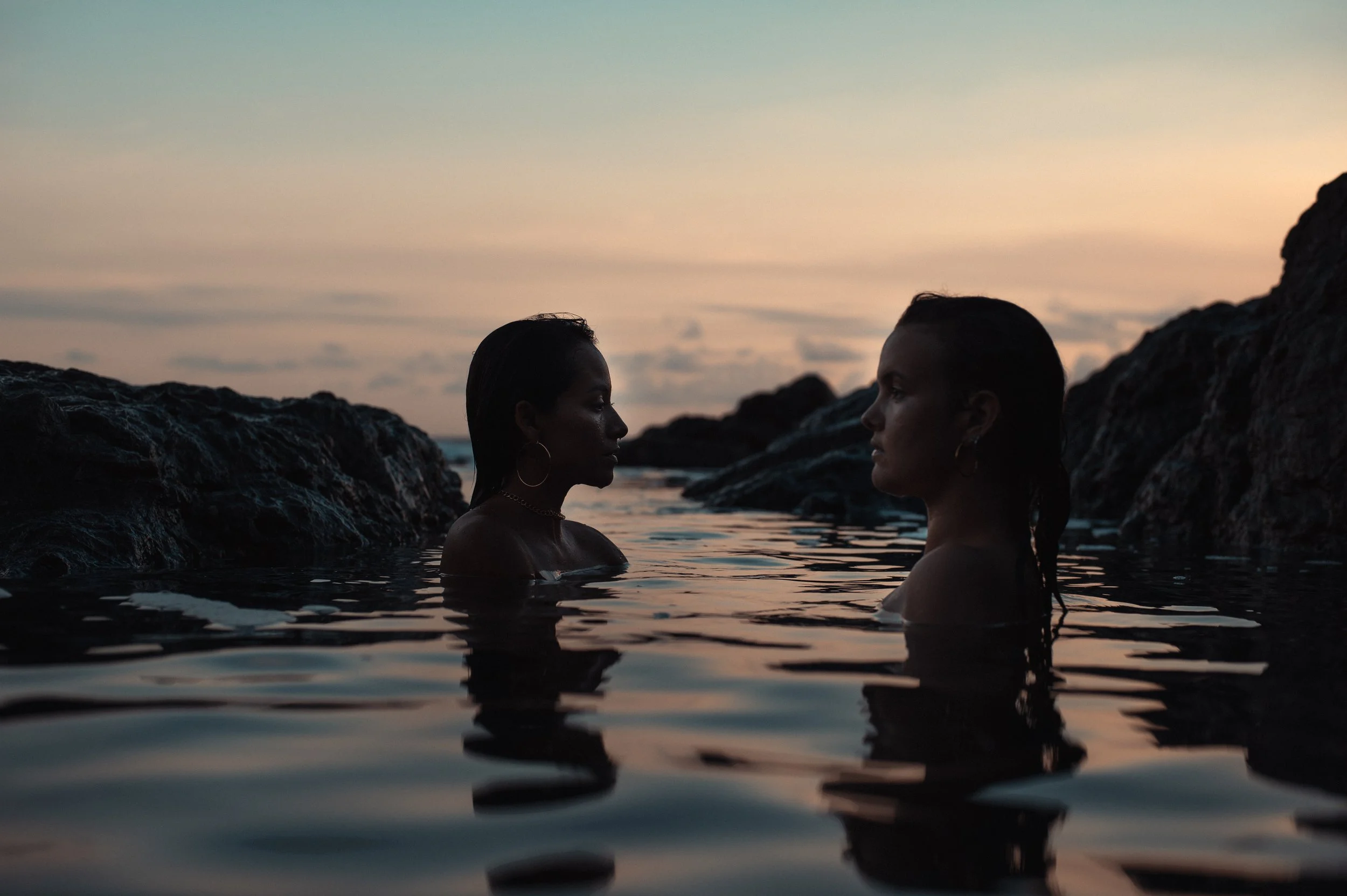 Two women with dark hair and hoop earrings are standing in the ocean at sunset, facing each other with rocky formations in the background.
