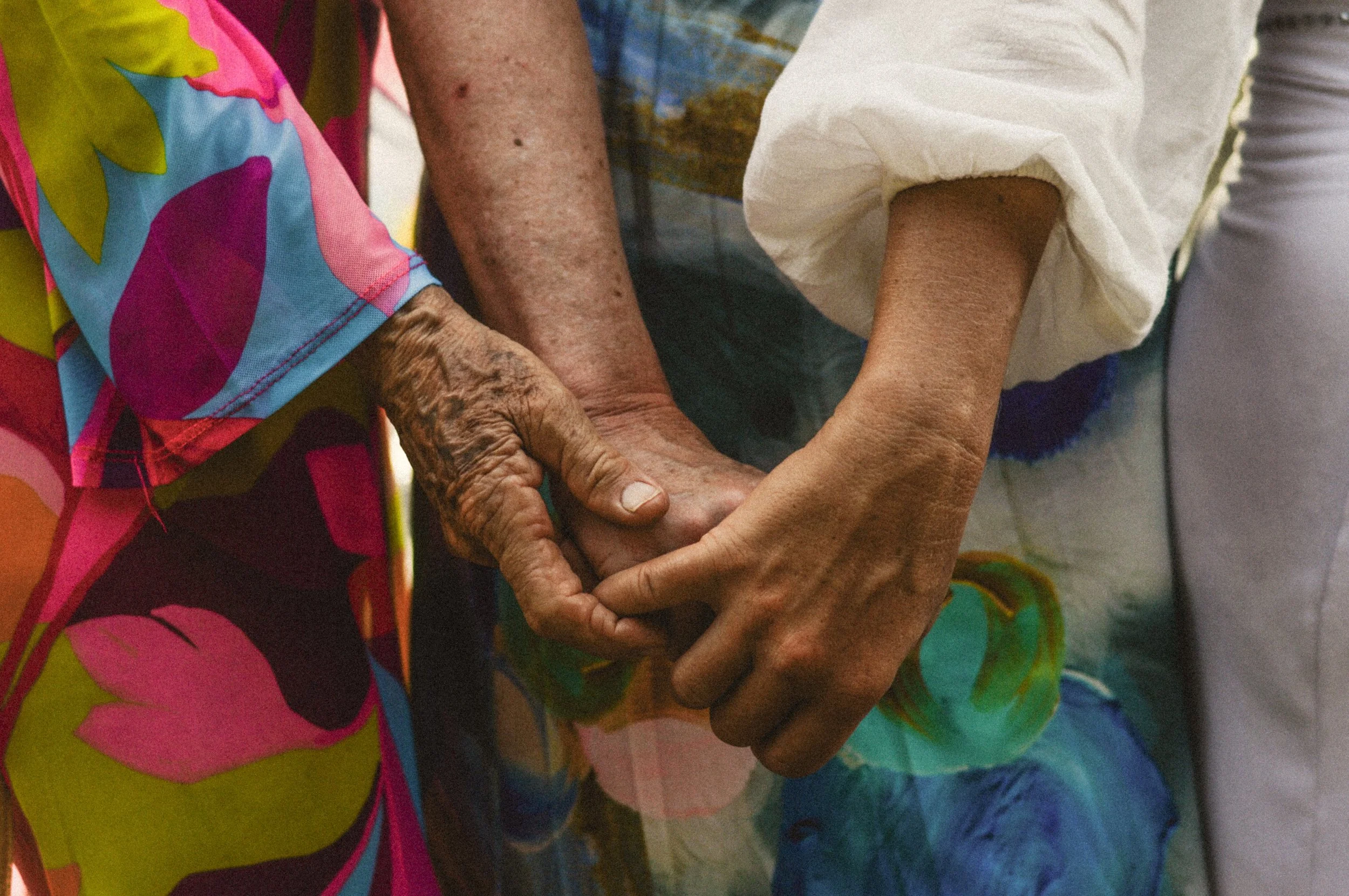 Close-up of elderly hands holding a younger person's hand, with colorful clothing in the background.