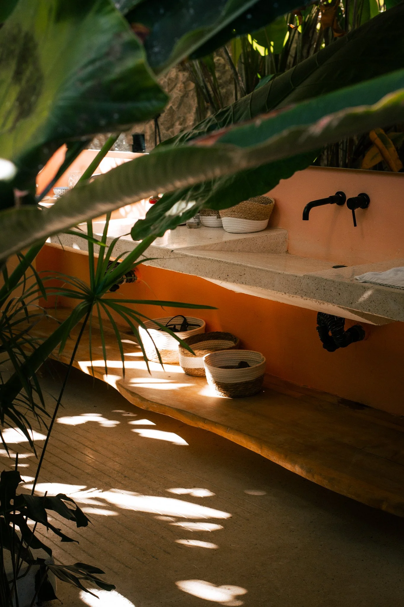 A rustic kitchen space with a concrete countertop, black faucet, and various woven baskets. Large plant leaves partially obscure the view, and warm sunlight creates patterned shadows on the wooden floor.