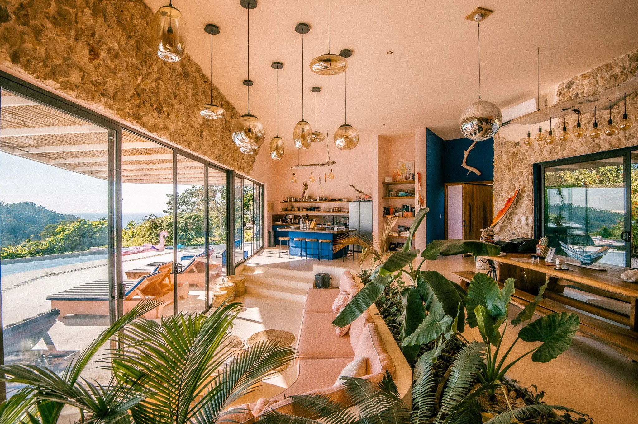 A spacious living room with large glass sliding doors leading to an outdoor patio with pool and lounge chairs. Inside, there is a pink sofa surrounded by green plants, a wooden dining table, and ceiling lights, with a stone accent wall and a disco ba