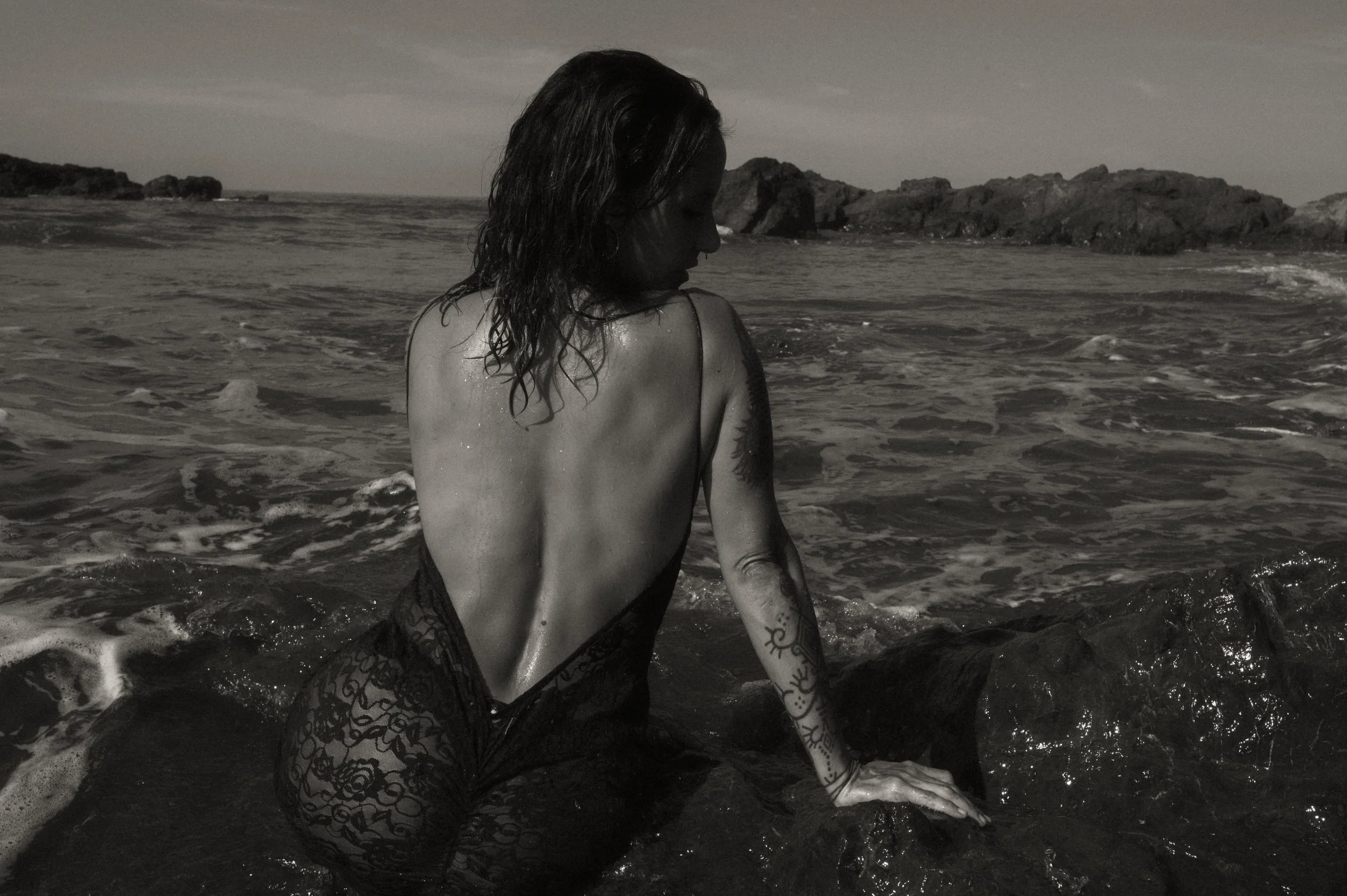 A woman with wet hair and tattoos on her arm sitting in the ocean, facing away, with rocks and cloudy sky in the background, in black and white.