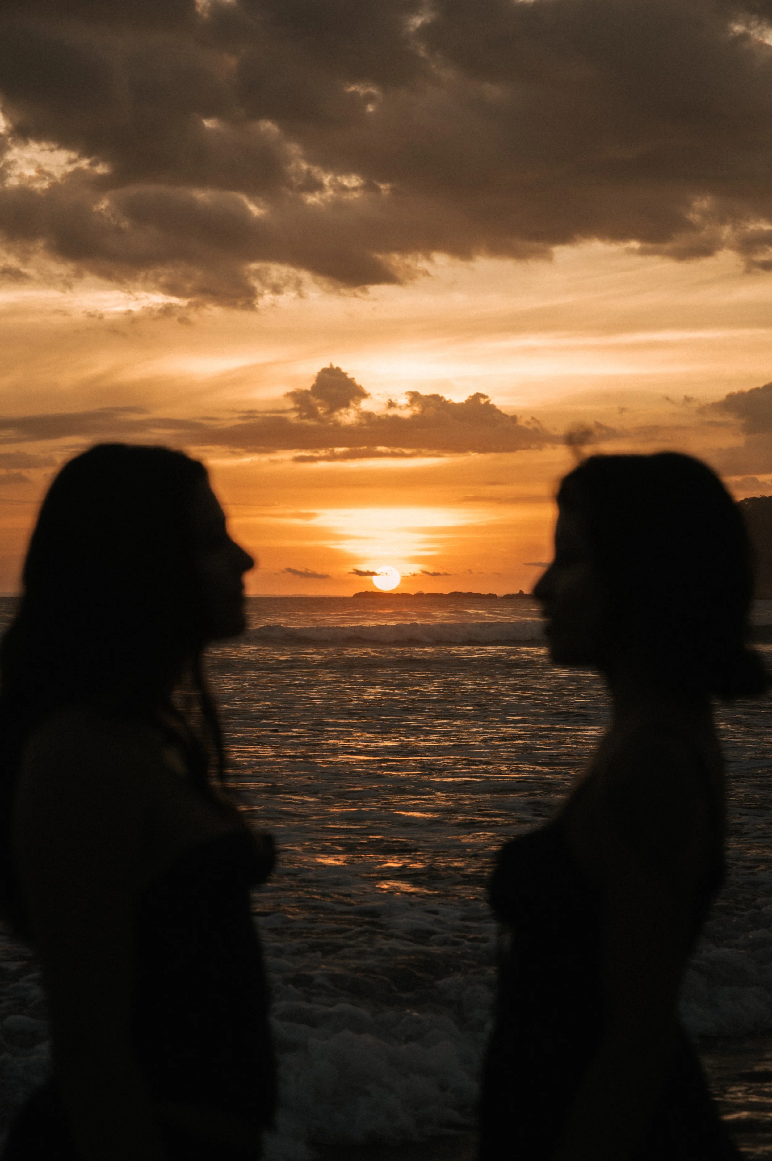 Silhouette of two women facing each other on the beach with a sunset sky and ocean in the background.