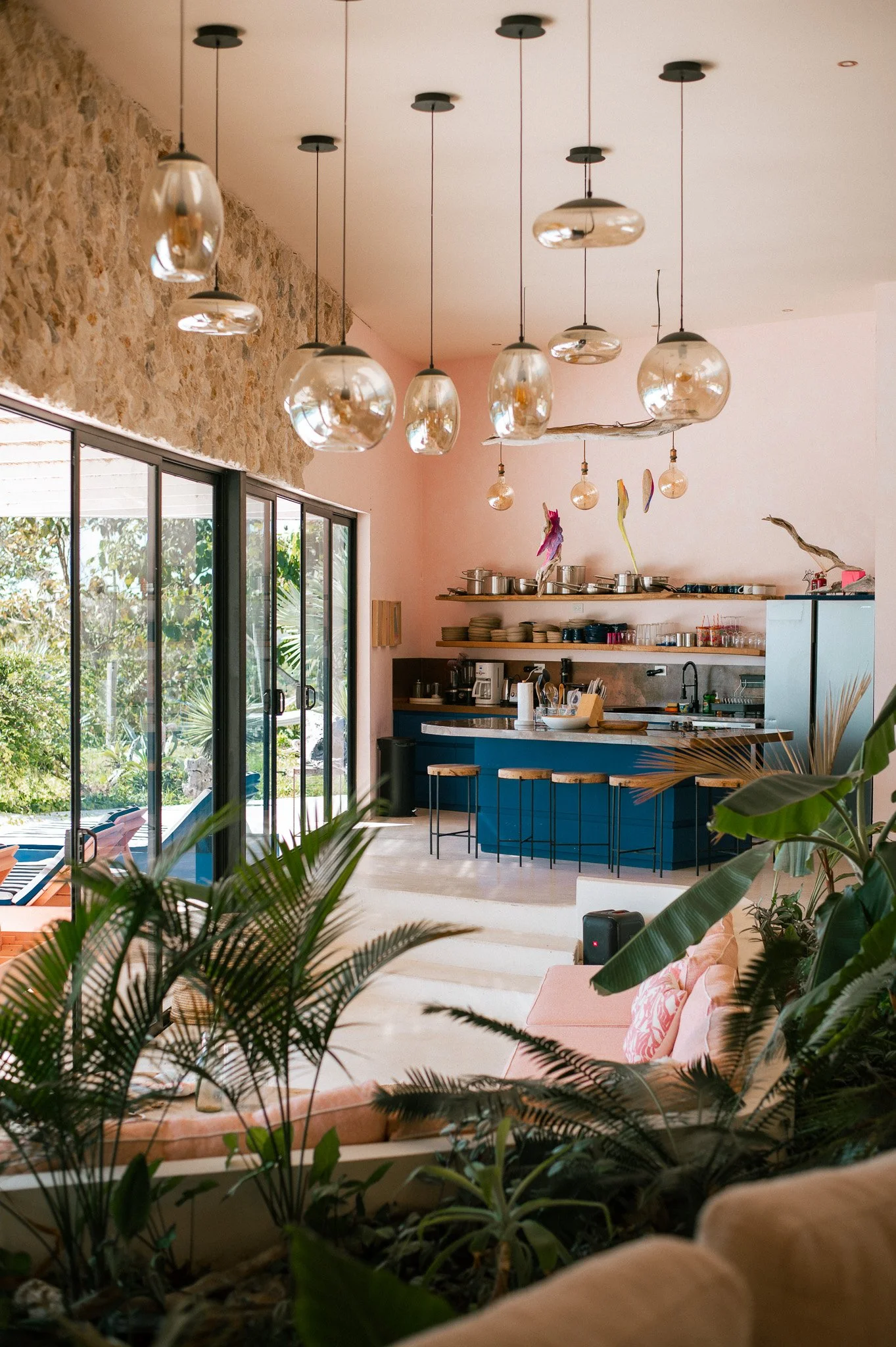 Interior of a modern kitchen and living area with large glass sliding doors, hanging pendant lights, open shelves with dishes, plants in the foreground, and a pastel pink and blue color scheme.