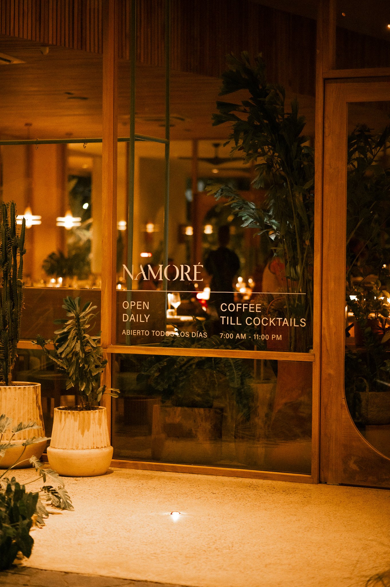 View of a warmly lit restaurant interior through glass doors with wooden framing, showing plants and seating inside, with restaurant hours and name displayed on the door.
