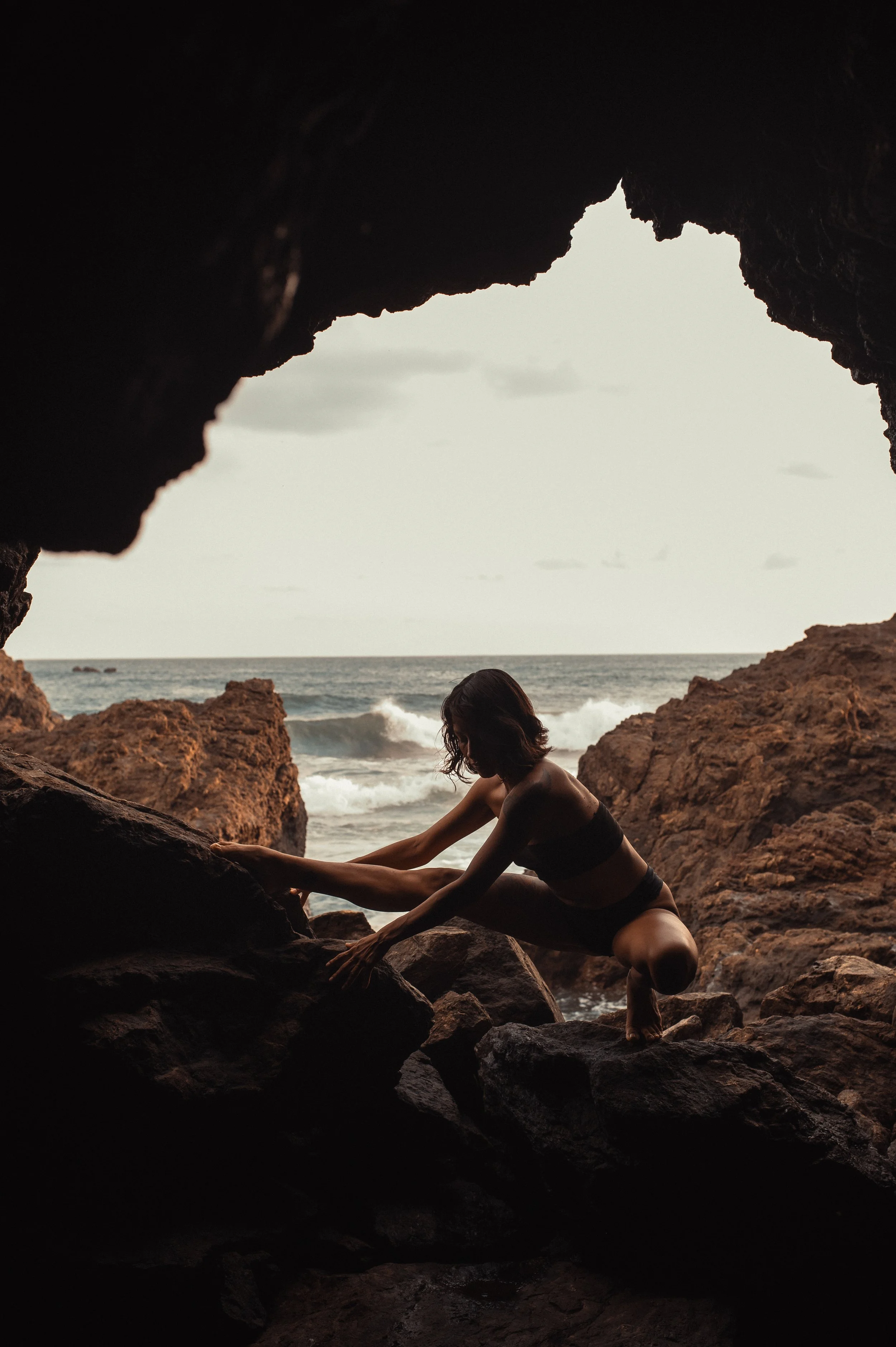 A woman practicing yoga on rocks near the ocean, seen through a cave opening with the ocean in the background.