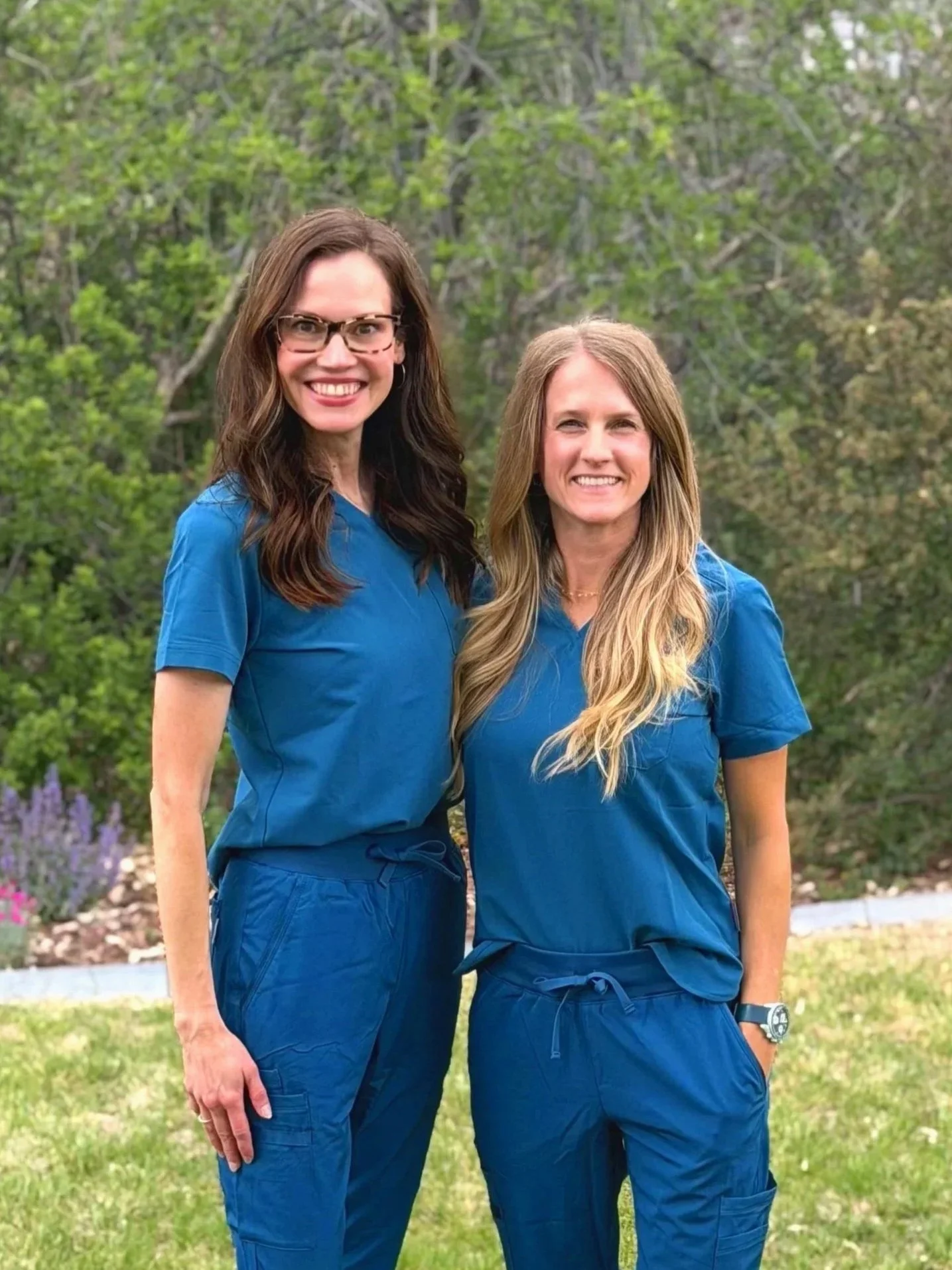 Two women in blue scrubs standing outdoors with greenery in the background, smiling at the camera.