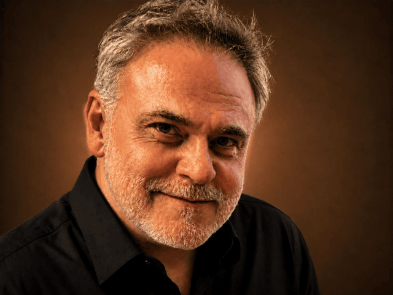 Close-up photograph of a middle-aged man with gray hair, a beard, and a black shirt, smiling slightly with a brown background.