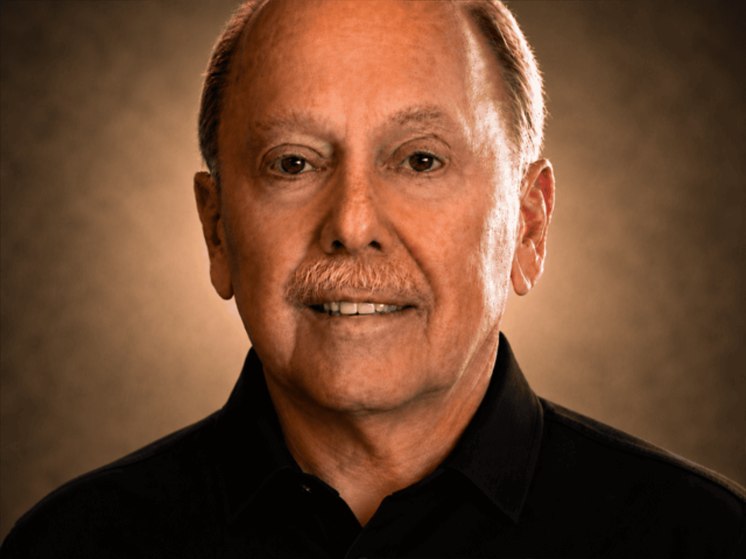 Close-up portrait of an older man with light skin, gray hair, and a mustache, wearing a black shirt, with a warm brown background.