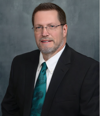 A professional man with glasses, a beard, and short brown hair wearing a dark suit and teal tie, posing against a neutral background.