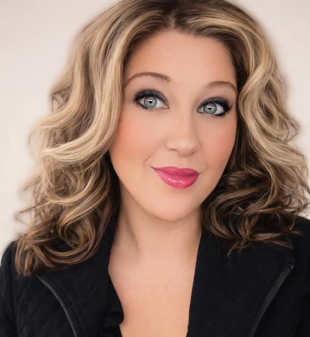 Close-up portrait of a woman with shoulder-length, wavy blonde hair, blue eyes, and pink lipstick, wearing a dark top.