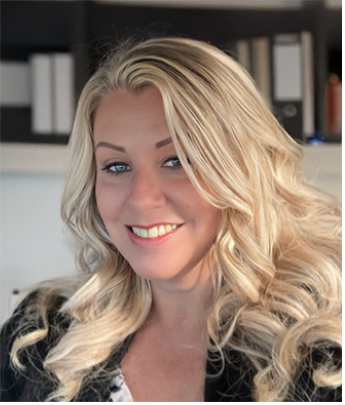 A blonde woman with curly hair smiling at the camera, in an indoor setting with books and shelves in the background.
