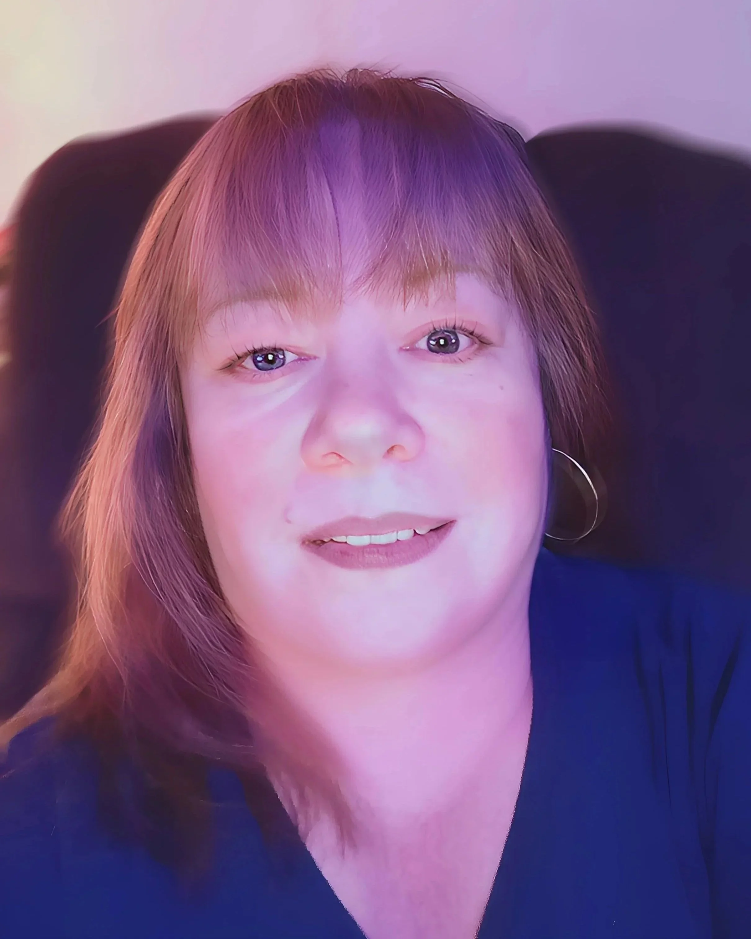 Close-up portrait of a woman with shoulder-length light brown hair, purple highlights, blue eyes, and wearing hoop earrings, smiling slightly, against a dark background.