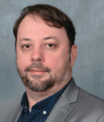 Headshot of a man with dark hair and a beard wearing a gray blazer and a dark blue dotted shirt, with a neutral gray background.