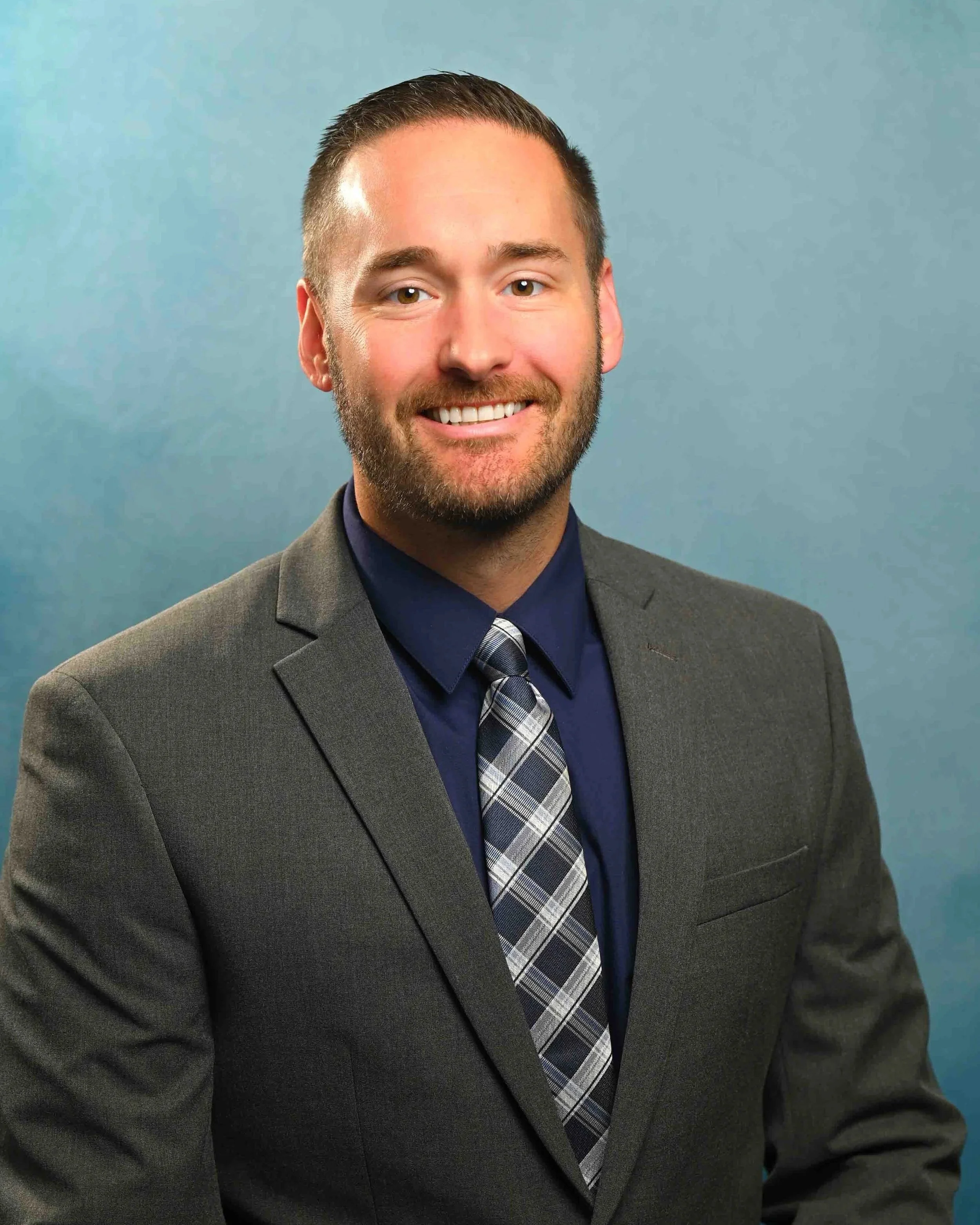 Professional man in a dark gray suit, navy shirt, and plaid tie, smiling against a muted blue background.