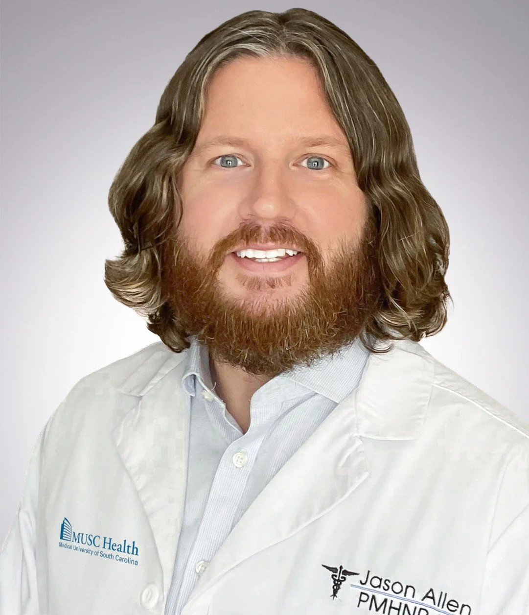 A man with long brown hair, a beard, and blue eyes, wearing a white medical coat with "MUSC Health" and "Jason Allen PMHNP" embroidered on it, smiling against a plain gray background.