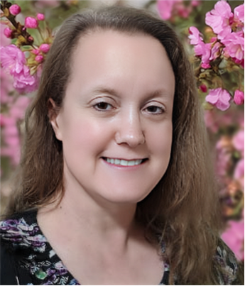 A woman with long brown hair smiling in front of pink flowers.