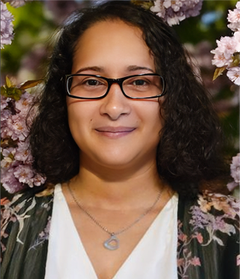 Portrait of a woman with glasses and curly dark hair, standing among pink cherry blossoms, smiling gently.