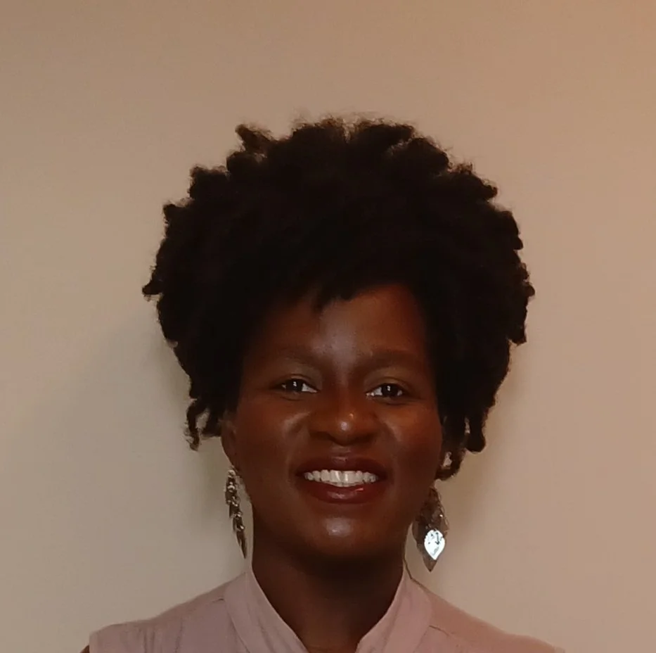 A smiling woman with dark, natural curly hair wearing silver earrings and a light-colored top against a plain wall.