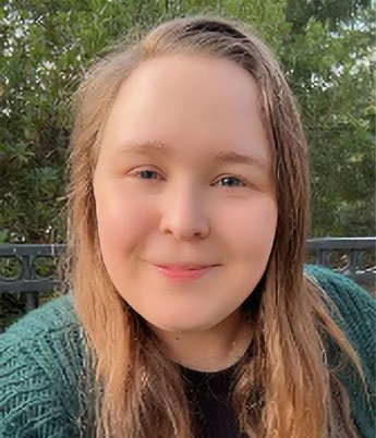 A young woman with long light brown hair smiling outdoors with greenery and a black metal fence in the background.