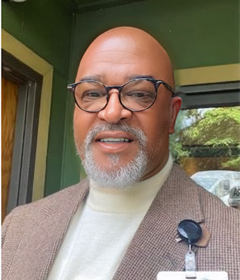 Close-up of a bald man with glasses and a beard, wearing a beige blazer over a cream turtleneck, standing indoors near a window with a green wall in the background.