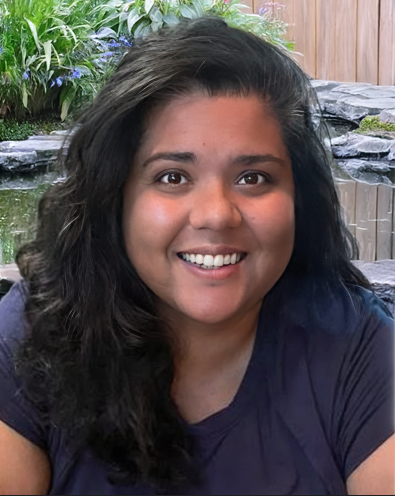 A woman smiling with long dark hair, outdoors near a water feature and greenery.