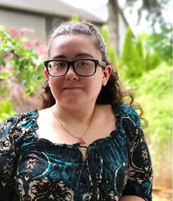 A young woman with glasses and curly hair, wearing a patterned blue and black dress, standing outdoors with greenery in the background.
