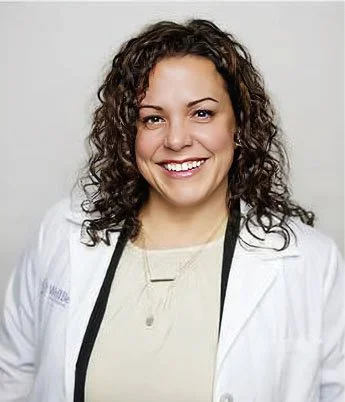 A woman with curly hair smiling, wearing a white lab coat and light-colored shirt, against a plain background.