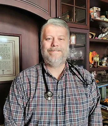 A man with gray hair and a beard smiling, wearing a plaid shirt and a stethoscope around his neck, standing in front of a wooden bookcase filled with books and decorative items.