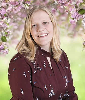 Smiling woman with blonde hair standing outdoors among pink flowering trees.