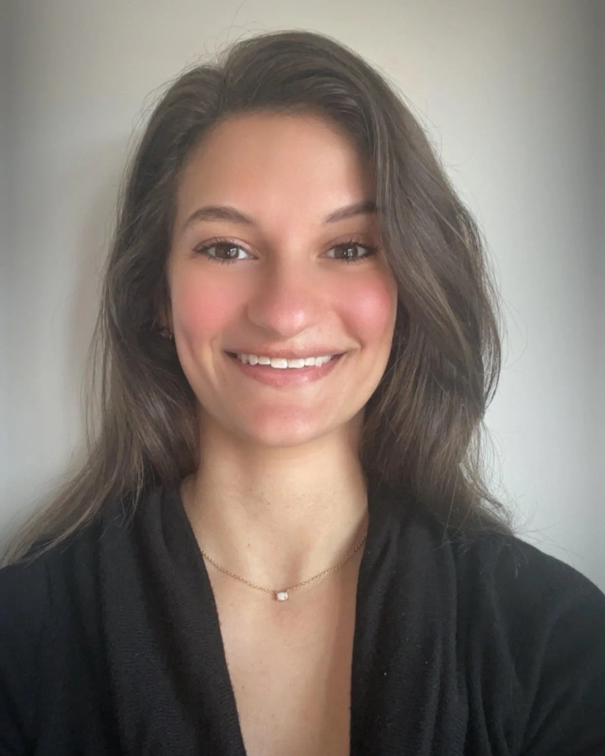 A woman with long, wavy brown hair, smiling, wearing a black shirt and a delicate gold necklace with a small square pendant.