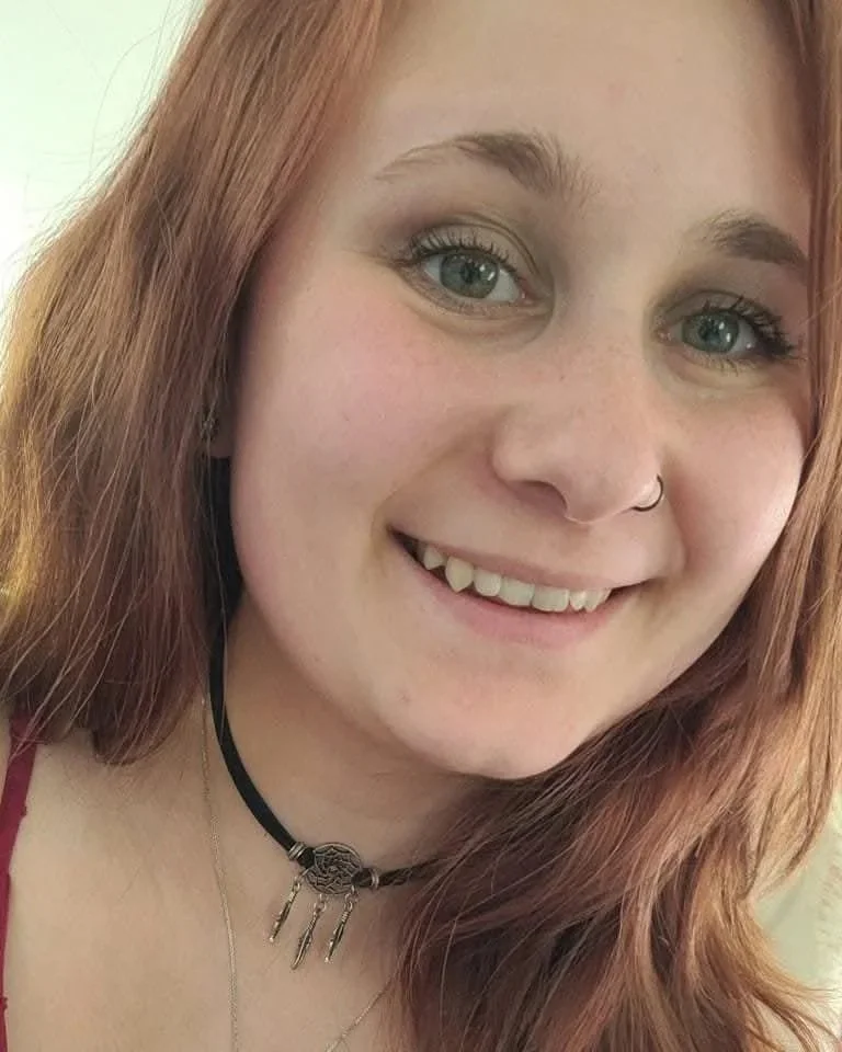 A young woman with red hair smiling, showing her teeth with a small gap, wearing a nose ring and a black choker with a pendant and small dangling charms.