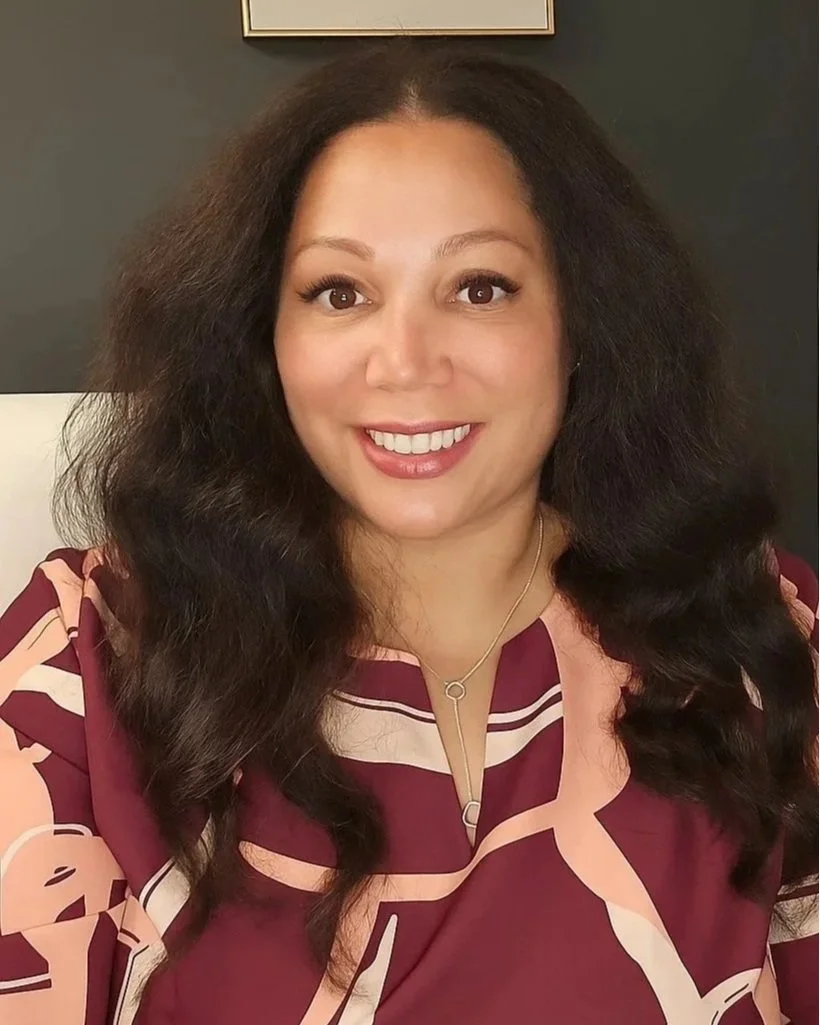A woman with long, dark, voluminous hair smiling at the camera, wearing a patterned burgundy and cream top, and a delicate necklace, with a dark wall and a framed picture in the background.