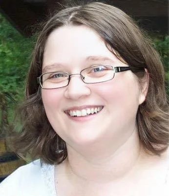A woman with glasses and shoulder-length brown hair smiling outdoors.