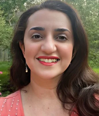 A woman with long brown hair smiling outdoors with trees in the background.