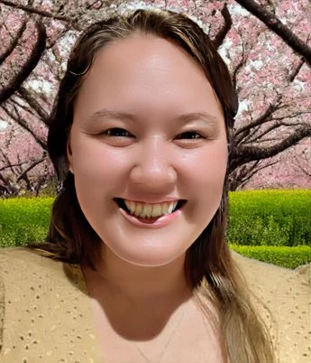 Smiling woman with shoulder-length brown hair, standing outdoors in front of blooming cherry blossom trees.
