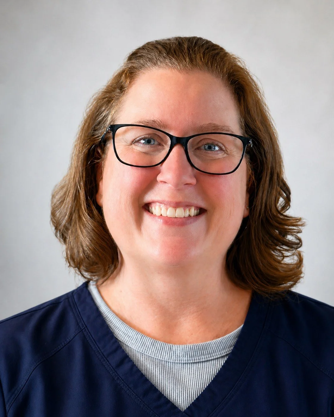 A woman with glasses smiling, wearing a navy blue medical scrubs, and standing against a gray background.