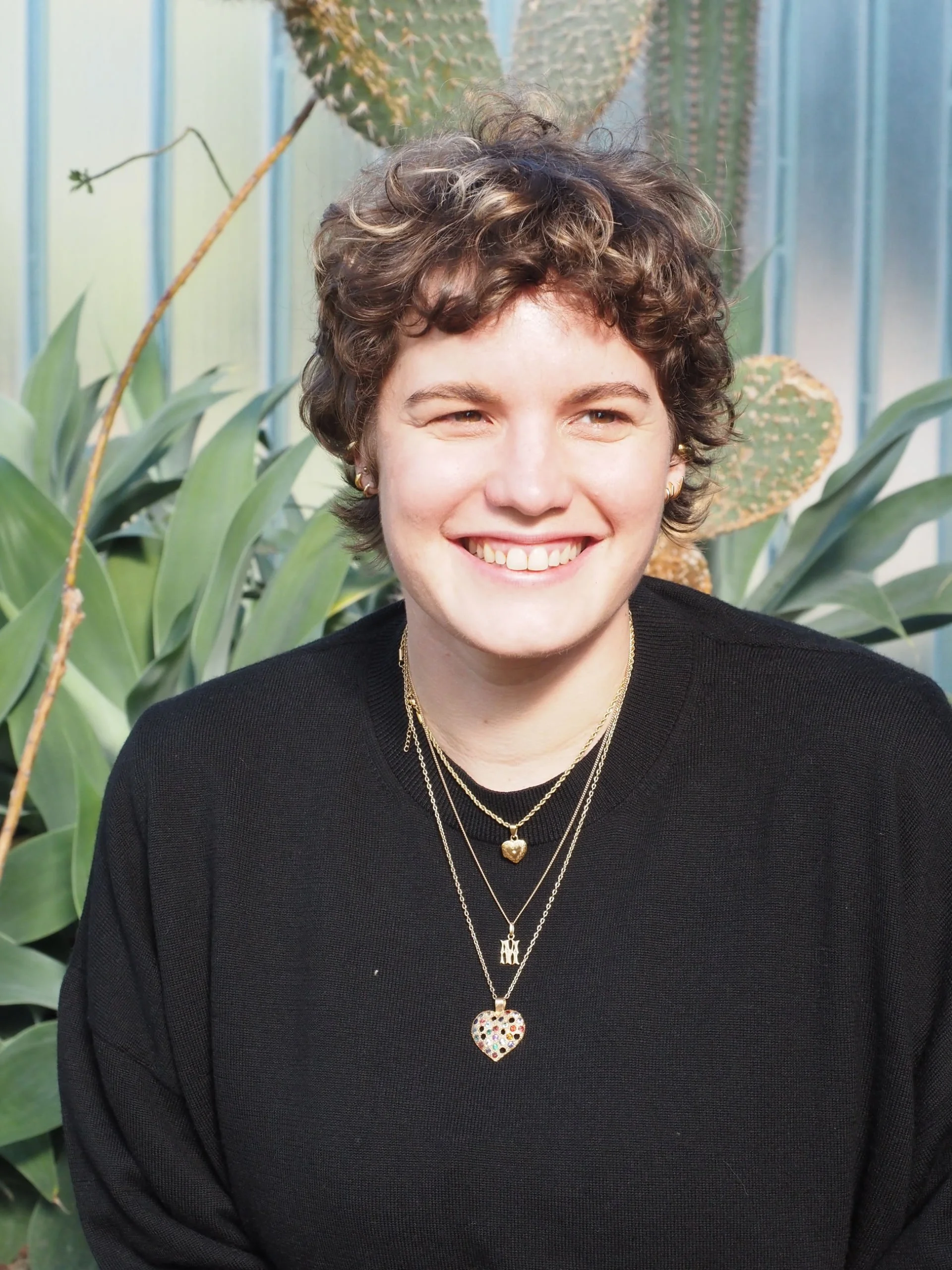 Une jeune femme souriante avec des cheveux bouclés courts, portant une chemise noire et plusieurs colliers, dont un avec un pendentif en forme de cœur en métal avec des pierres colorées, devant un fond de plantes vertes dont des cactus.