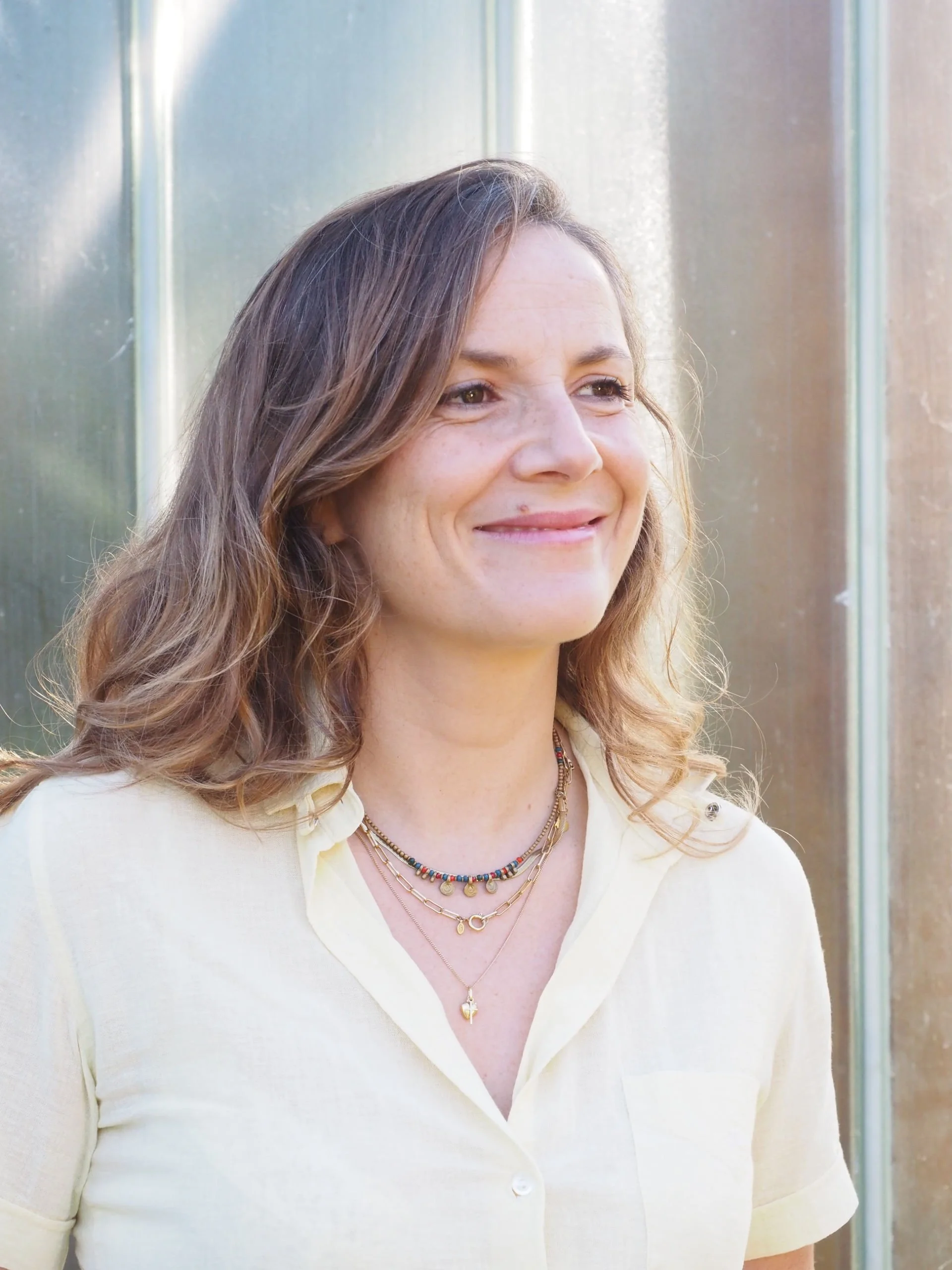 Portrait d'une femme souriante portant des bijoux, en chemise claire, devant un fond métallique.