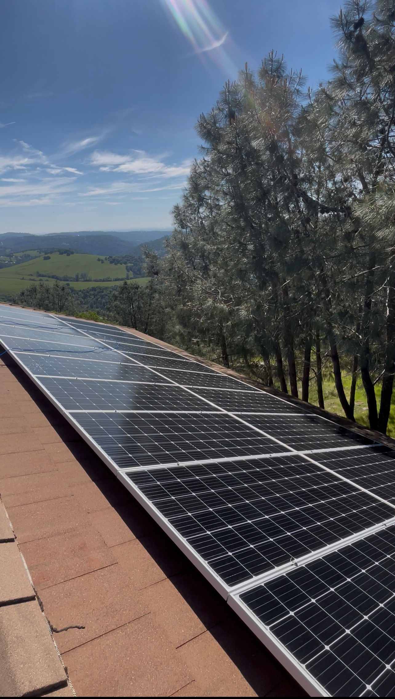 Solar panels on a rooftop overlooking a hilly landscape with green fields, trees, and a clear blue sky with clouds and lens flare.