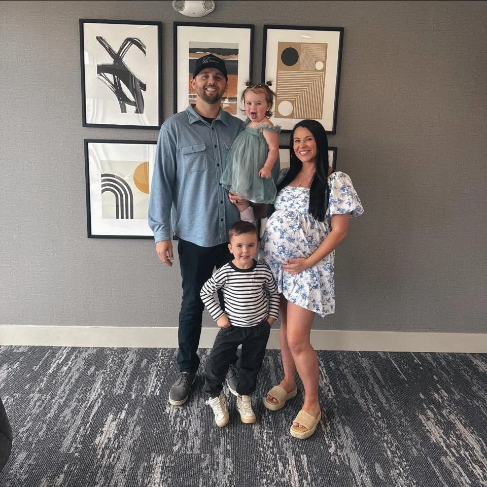 Family photo of a man, woman, and two children standing against a gray wall with abstract art frames. The woman is pregnant and smiling, the man is wearing a cap and a button-up shirt, the girl is being held and is smiling widely, and the boy is standing in front with his hand on his hip.