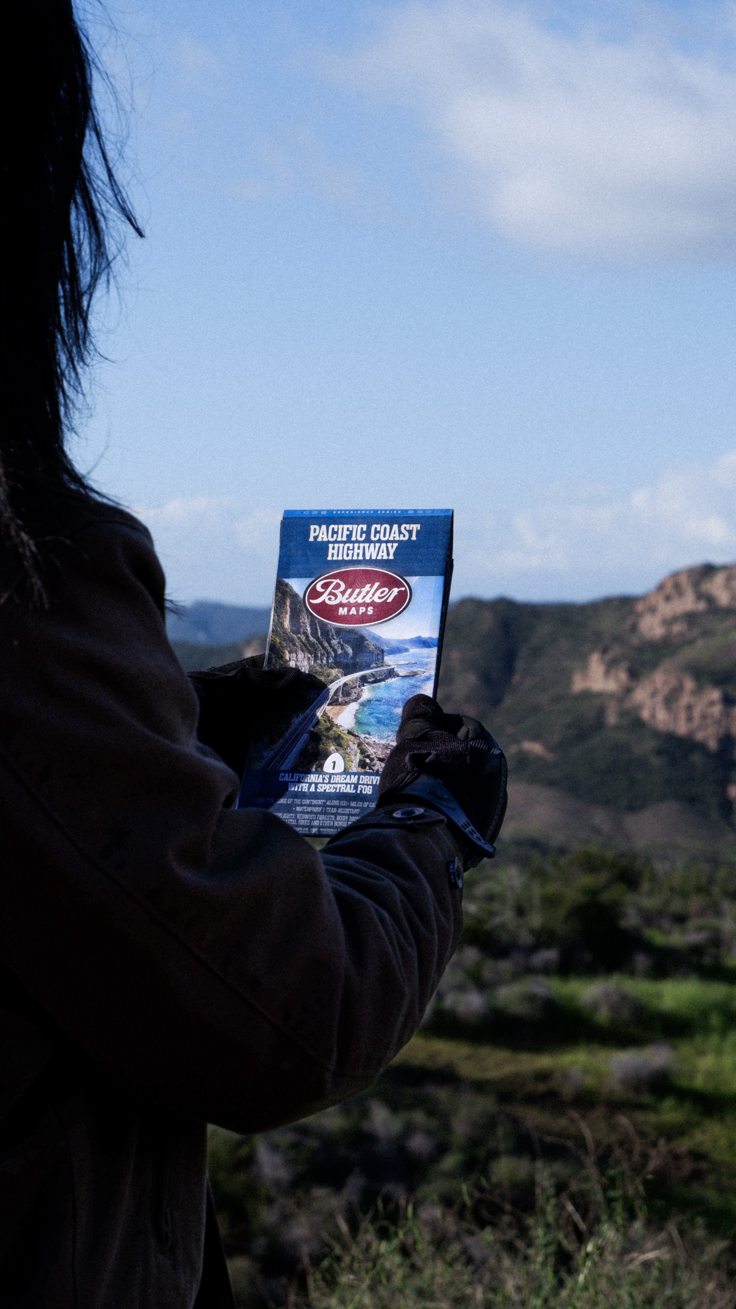 Motorcyclist holding a Butler Maps Pacific Coast Highway map overlooking scenic California coastal mountains.
