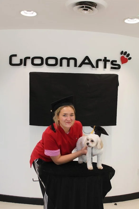 Young woman in graduation cap and gown with a puppy on a table in front of a GroomArts sign.